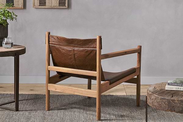 The Dalibar Leather Lounger - Tan by nkuku, crafted from brown buffalo leather with a natural acacia wood frame, is shown from the back on a textured gray rug beside a side table with a glass of water and another table with books.