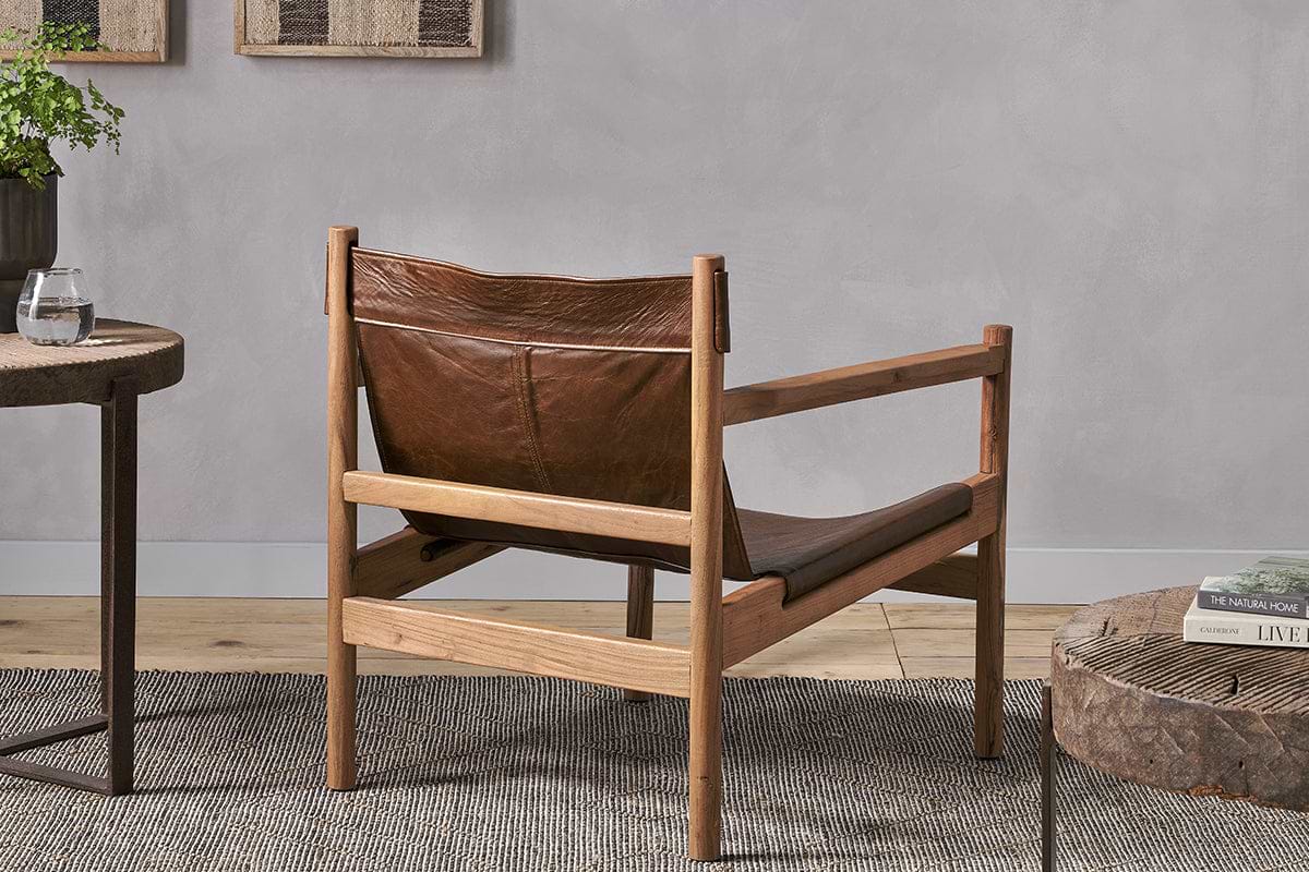 The Dalibar Leather Lounger - Tan by nkuku, crafted from brown buffalo leather with a natural acacia wood frame, is shown from the back on a textured gray rug beside a side table with a glass of water and another table with books.