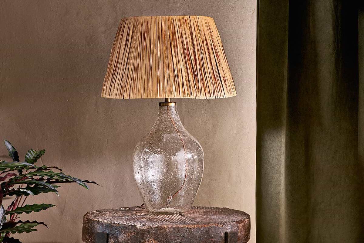 The nkuku Damini Recycled Glass Table Lamp, featuring a textured clear base and woven shade, is placed on a rustic wooden table beside a leafy plant, with a dark green curtain against a beige wall.