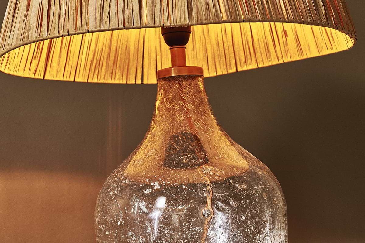 A close-up of the nkuku Damini Recycled Glass Table Lamp, featuring a textured, recycled glass base and pleated beige shade, glowing warmly and casting soft light against a neutral wall.