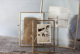 The Danta Antique Brass Frames by nkuku display vintage photos and a faded note. Set on a textured surface with a blurred background, the brass frames evoke nostalgic, timeless elegance.