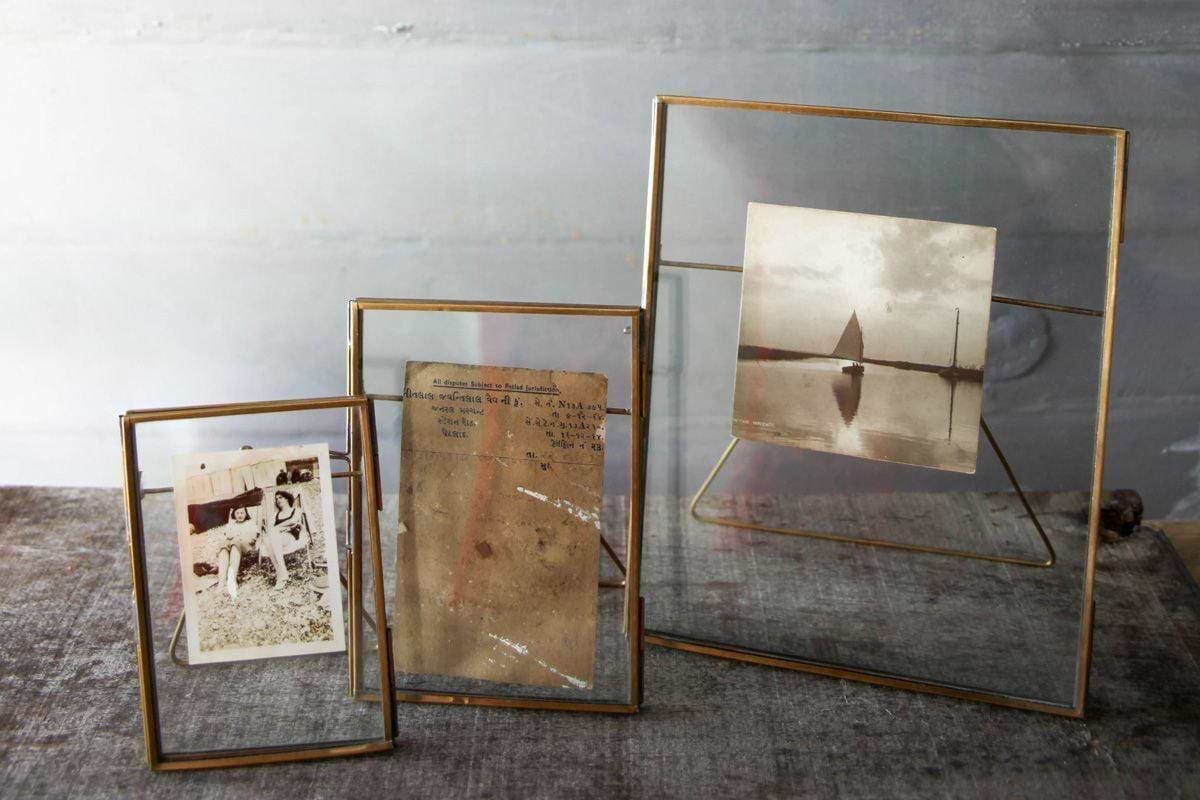 Three elegant Danta Antique Brass Frames by nkuku of varying sizes stand on a textured surface. The left displays a sepia photo of a woman, the middle holds a faded note, and the right shows a vintage sailboat on calm water.