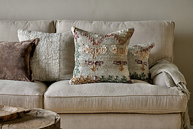 A beige sofa with three textured pillows—one featuring the Dantri Jute & Cotton Cushion Cover in Soft Grey by nkuku, a brown handloom cushion, and a pastel geometric design. A light throw blanket rests on the right armrest. The background wall is light gray; a rustic wooden table sits in front.