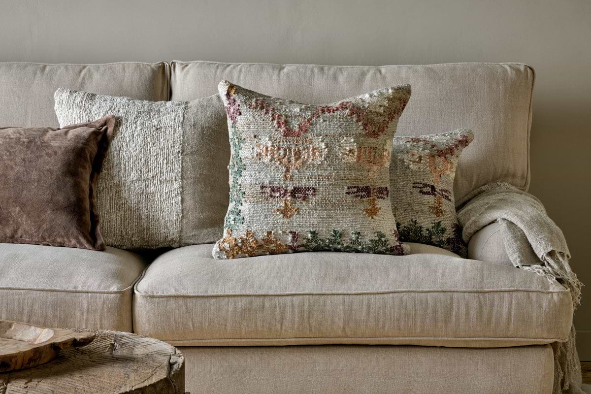A beige sofa with three textured pillows—one featuring the Dantri Jute & Cotton Cushion Cover in Soft Grey by nkuku, a brown handloom cushion, and a pastel geometric design. A light throw blanket rests on the right armrest. The background wall is light gray; a rustic wooden table sits in front.