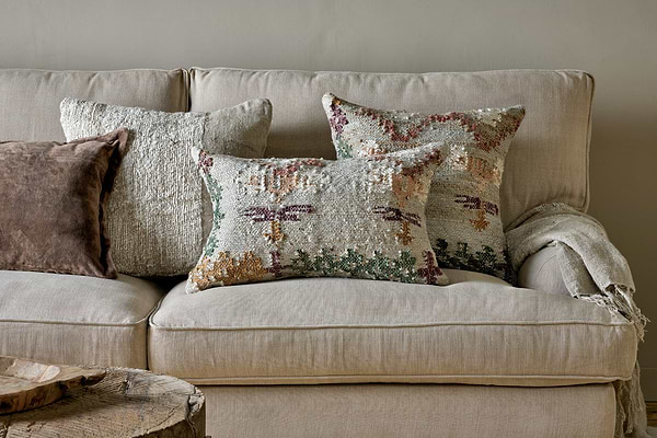 A beige fabric sofa features four textured throw pillows, including the nkuku Dantri Jute & Cotton Cushion Cover in soft grey, beneath a light woven blanket. A rustic wooden table sits in the foreground.