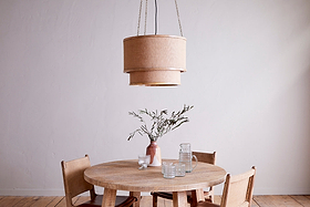 A round wooden table with four chairs sits on a light wood floor. Above, the nkuku Darat Mango Wood & Jute Pendant in Natural adds rustic charm. On the table are a pink vase with green branches, two clear glasses, and a clear pitcher.