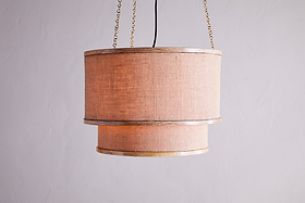 The Darat Mango Wood & Jute Pendant in Natural by nkuku features a double-layered, textured jute drum shade suspended from two gold chains with a visible black cord, casting a warm glow against a light gray wall.