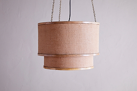 The Darat Mango Wood & Jute Pendant by nkuku features a natural jute shade with mango wood trim, suspended by a black cord and two brass chains, creating rustic elegance against a light gray wall.