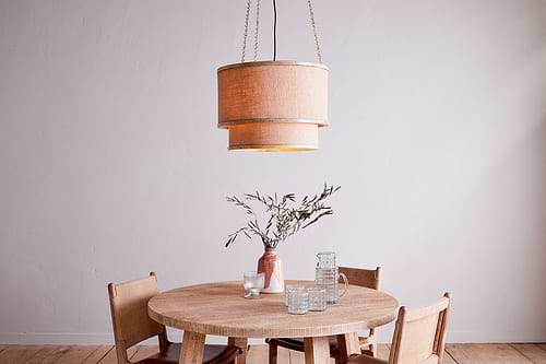 A round wooden table with four chairs surrounds a brown vase with greenery and clear glasses. Above, the nkuku Darat Mango Wood & Jute Pendant - Natural hangs, enhancing the minimalist room’s light walls and wooden floor.