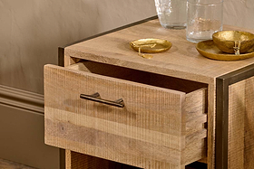 The Dasai Mango Wood Bedside Table by nkuku, featuring an open drawer with a simple metal handle, displays two gold bowls with jewelry and two clear glasses on top. Soft lighting highlights its handcrafted mango wood grain against a beige wall.