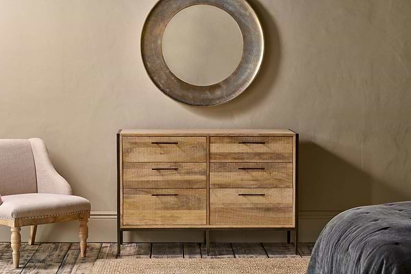 The Dasai Mango Wood Chest of Drawers by nkuku, featuring eight drawers, is set against a beige textured wall with a large round metallic mirror above. A light-upholstered chair, gray bed, and wooden floor create a cozy bedroom scene.