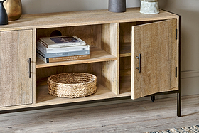 The Dasai Mango Wood Media Unit by nkuku features light mango wood with black handles, set on a wood floor. One door is open to show two shelves with stacked books and a woven basket. Decorative objects adorn this stylish console.