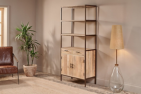 A modern living room showcases the nkuku Dasai Mango Wood Storage Unit, featuring drawers and cabinet doors for stylish storage. To the left sits a potted plant and brown leather chair; to the right is a glass-base floor lamp with beige shade.