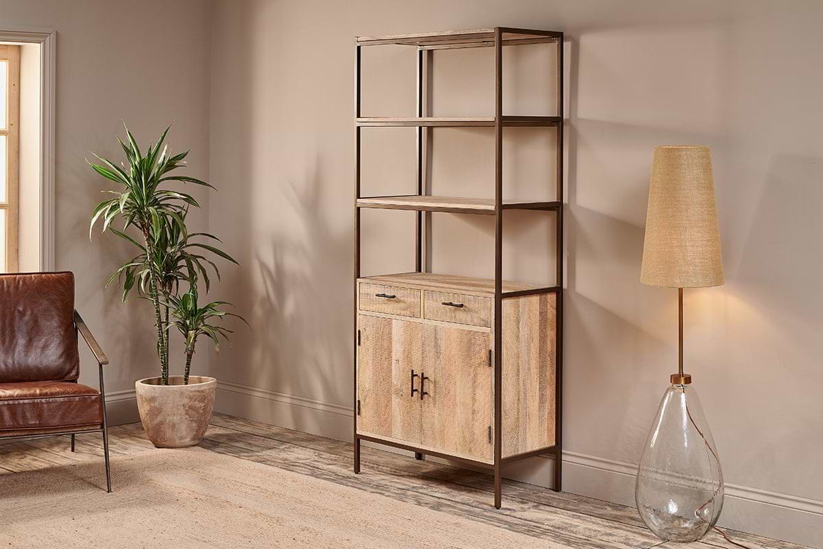 A modern living room showcases the nkuku Dasai Mango Wood Storage Unit, featuring drawers and cabinet doors for stylish storage. To the left sits a potted plant and brown leather chair; to the right is a glass-base floor lamp with beige shade.