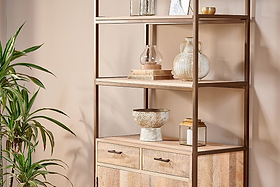 The Dasai Mango Wood Storage Unit by nkuku features metal accents and two drawers, styled with glass vases, a ceramic bowl, and candles. A leafy plant sits on the left against a beige wall for a chic, minimalist living room look.