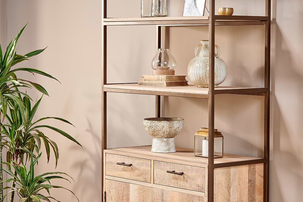 The Dasai Mango Wood Storage Unit by nkuku features metal accents and two drawers, styled with glass vases, a ceramic bowl, and candles. A leafy plant sits on the left against a beige wall for a chic, minimalist living room look.