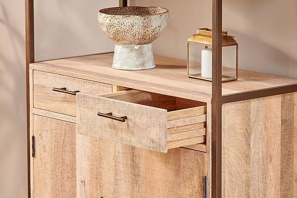 The nkuku Dasai Mango Wood Storage Unit features two doors and two drawers, one partially open. A speckled ceramic bowl and gold lantern with a white candle rest on top—stylish storage for your living room against a beige wall.