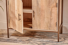 The Dasai Mango Wood Storage Unit by nkuku features two doors—one partially open showing an interior shelf—a natural mango wood grain finish, metal handles, slim metal legs, and stands on a textured wood-effect floor.