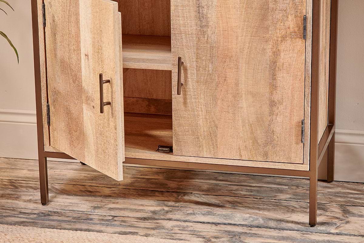 The Dasai Mango Wood Storage Unit by nkuku features two doors—one partially open showing an interior shelf—a natural mango wood grain finish, metal handles, slim metal legs, and stands on a textured wood-effect floor.