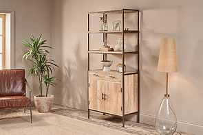 A modern living room includes the nkuku Dasai Mango Wood Storage Unit with decor, a tan glass-base floor lamp, potted plant, brown leather chair, and beige rug, all set against a light beige wall and wooden floor.