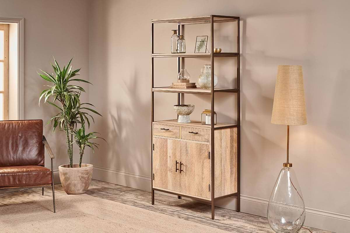 A modern living room includes the nkuku Dasai Mango Wood Storage Unit with decor, a tan glass-base floor lamp, potted plant, brown leather chair, and beige rug, all set against a light beige wall and wooden floor.