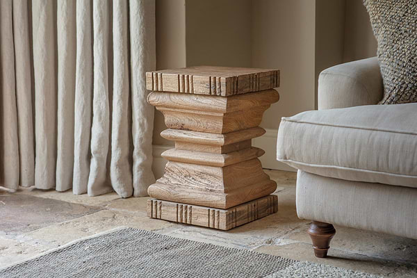 The Disha Carved Mango Wood Side Table by nkuku, with layered geometric details and natural grain, sits on a stone floor beside a beige armchair and cream curtains, with a textured rug nearby.