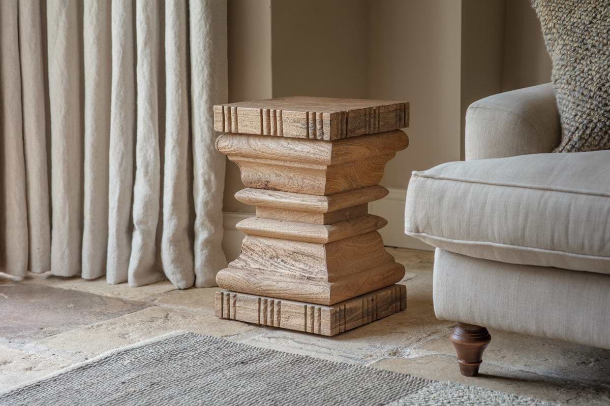 The Disha Carved Mango Wood Side Table by nkuku, with layered geometric details and natural grain, sits on a stone floor beside a beige armchair and cream curtains, with a textured rug nearby.