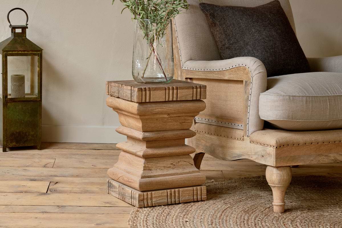 A rustic living room with a wooden floor, beige armchair with dark cushion, nkuku’s Disha Carved Mango Wood Side Table holding a clear vase with green branches, round woven rug, and vintage green lantern by the wall.