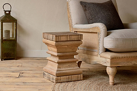 The nkuku Disha Carved Mango Wood Side Table - Natural, featuring a geometric layered design, stands beside a beige armchair with a dark cushion on a wooden floor, accompanied by a vintage green lantern against an off-white wall.