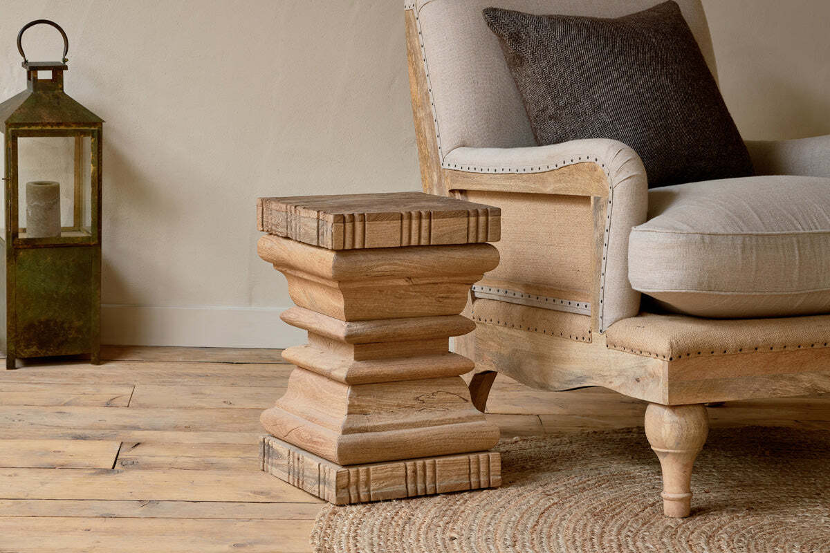 The nkuku Disha Carved Mango Wood Side Table - Natural, featuring a geometric layered design, stands beside a beige armchair with a dark cushion on a wooden floor, accompanied by a vintage green lantern against an off-white wall.