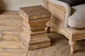 The Disha Carved Mango Wood Side Table by nkuku stands beside a light wood sofa with beige upholstery, creating a cozy, earthy vibe on a rustic floor next to a woven round rug.