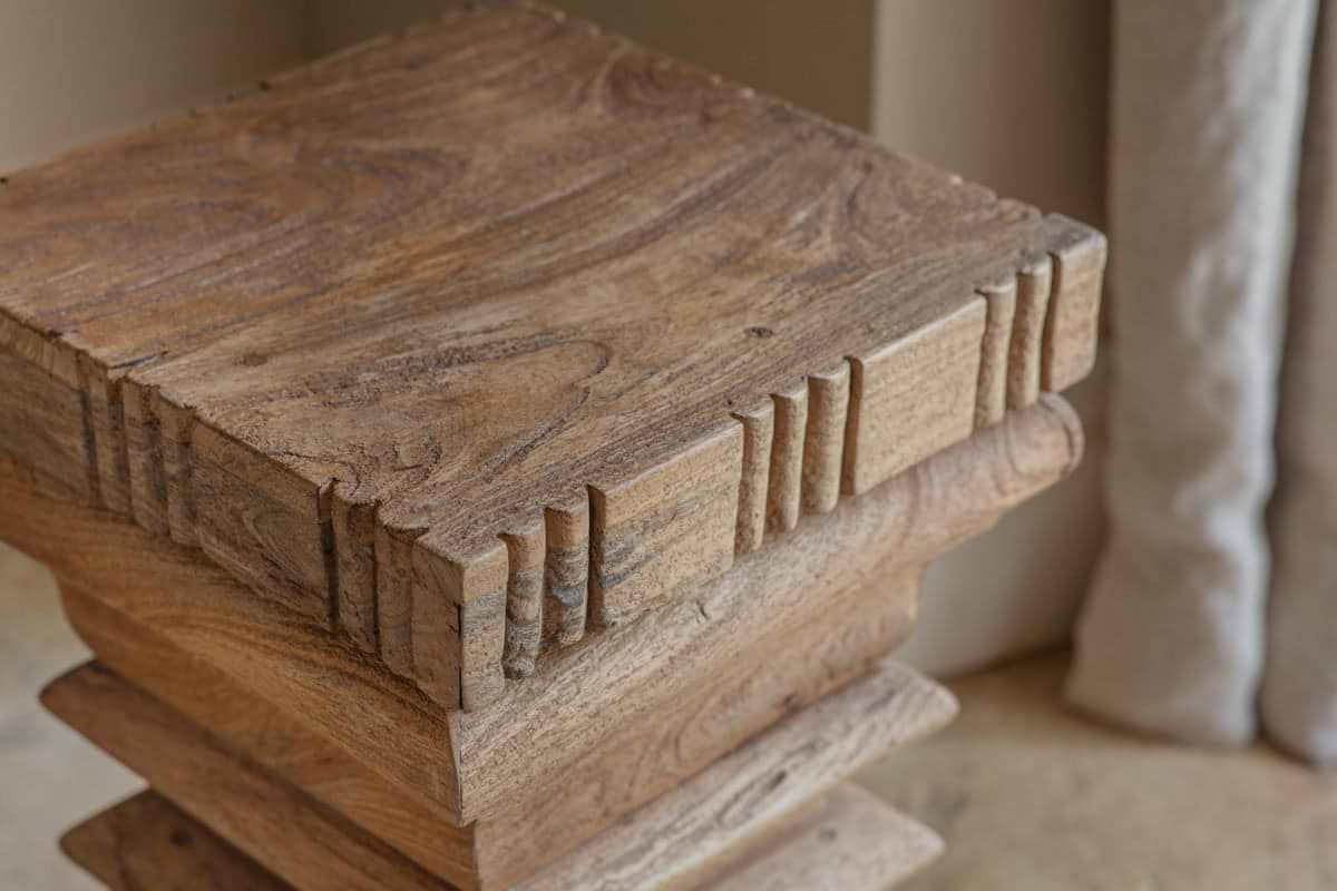 Close-up of the nkuku Disha Carved Mango Wood Side Table - Natural, featuring a square shape with hand-carved, notched edges and a natural finish that highlights the wood’s knots and grain patterns. Set against a neutral background with soft lighting.