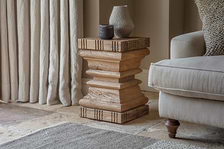 The nkuku Disha Carved Mango Wood Side Table in Natural stands by a beige sofa with a textured cushion, topped with two vases and a dark bowl. Stone flooring, cream curtains, and a woven rug complete the cozy rustic look.