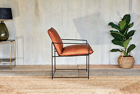 The Durium Leather Lounger - Aged Tan by nkuku, featuring a modern iron frame, rests on a woven rug. To the left sits a glass lamp on a gold table; to the right is a potted plant in a basket, set against a textured wall and wooden floor.
