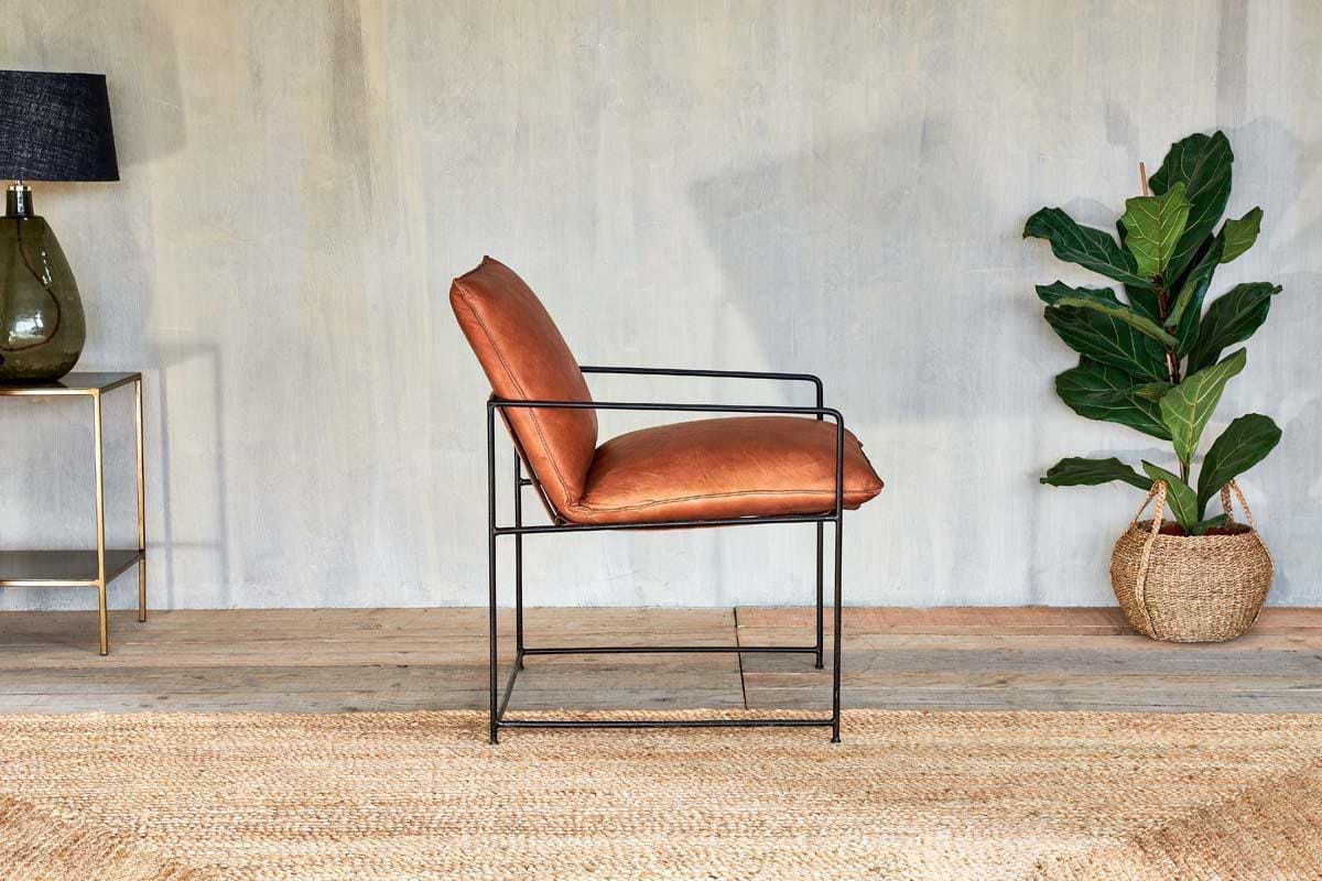 The Durium Leather Lounger - Aged Tan by nkuku, featuring a modern iron frame, rests on a woven rug. To the left sits a glass lamp on a gold table; to the right is a potted plant in a basket, set against a textured wall and wooden floor.