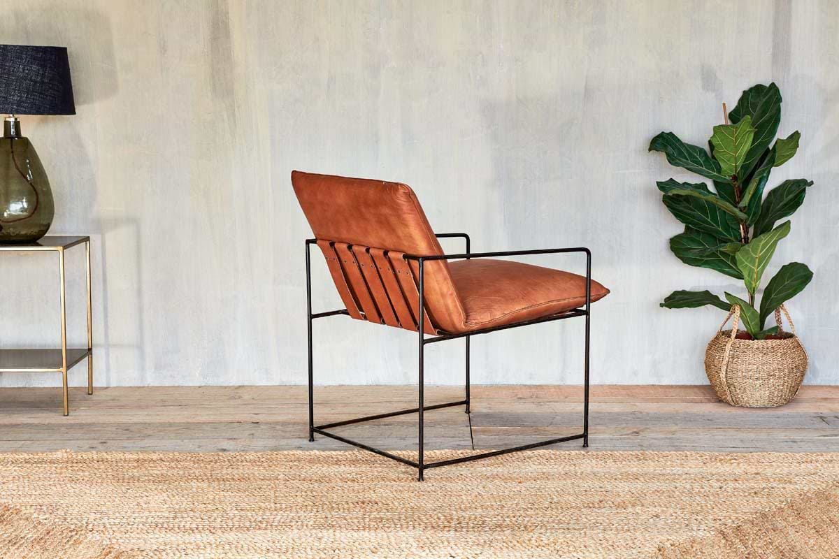 A modern room centers on the nkuku Durium Leather Lounger - Aged Tan with an industrial iron frame atop a woven rug. A glass lamp on a side table sits left, while a leafy plant in a woven basket stands to the right against a light wall.