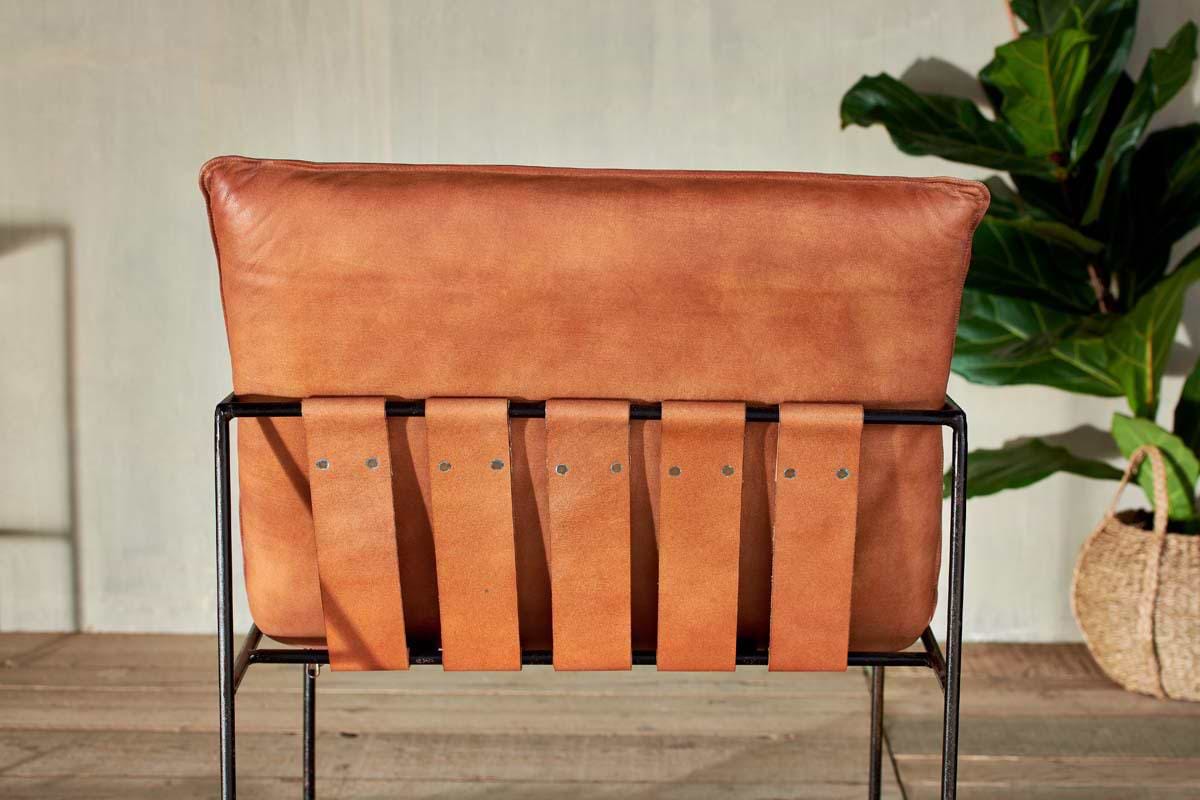 The Durium Leather Lounger - Aged Tan by nkuku is shown from the back, with wide leather straps on an iron frame. A green plant and woven basket sit on a wooden floor beside a light-colored wall.