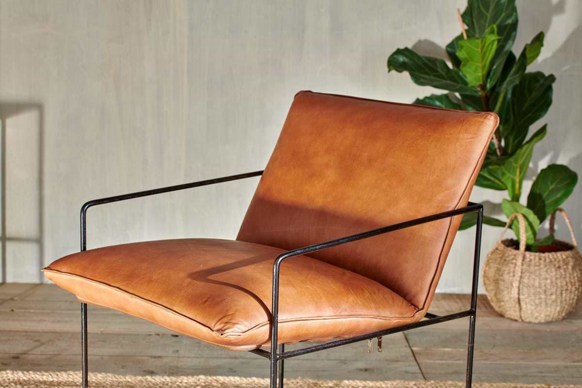 The nkuku Durium Leather Lounger - Aged Tan features a slim black iron frame and tan leather seat, set on a rug with a potted plant placed behind it.