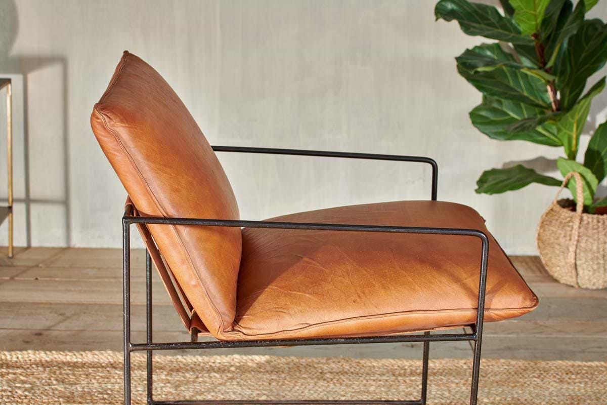 The nkuku Durium Leather Lounger - Aged Tan features a caramel brown leather seat and industrial iron frame, set on a woven rug with a green potted plant in the background against a light textured wall.