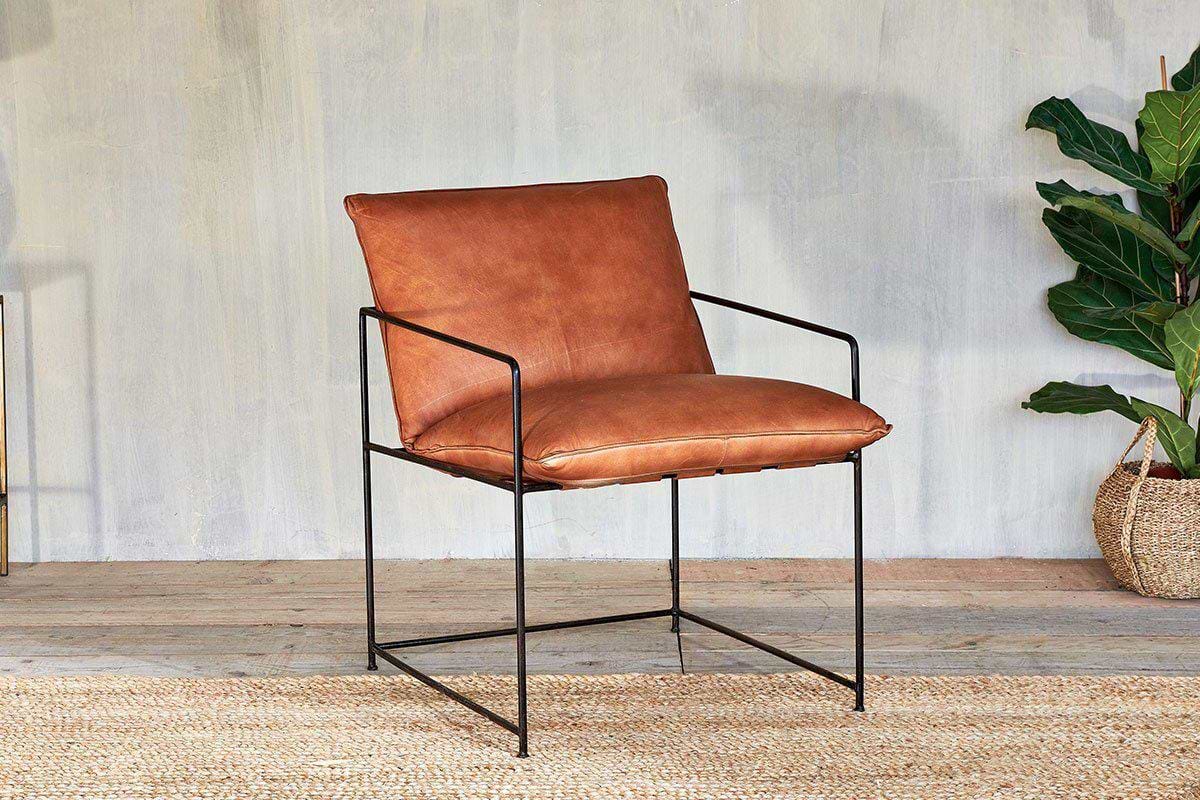 The nkuku Durium Leather Lounger - Aged Tan features a brown leather seat and backrest with slim black iron framework, placed on a woven rug. A green potted plant in a basket and a light gray textured wall add the finishing touches.
