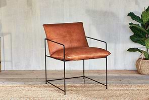 The nkuku Durium Leather Lounger - Aged Tan features a brown leather seat and backrest with slim black iron framework, placed on a woven rug. A green potted plant in a basket and a light gray textured wall add the finishing touches.