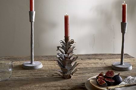 A rustic wooden table features two metal candlesticks, including the nkuku Elagalu Pine Cone Candle Holder in zinc. To the right is a wooden plate with sliced figs and cutlery, while a clear glass of water sits to the left against a plain wall.