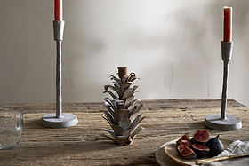 A rustic whitewashed wooden table displays two gray candlesticks with red candles, the nkuku Elagalu Pine Cone Candle Holder in zinc, a glass of water, and a wooden plate of figs against a neutral minimal background.