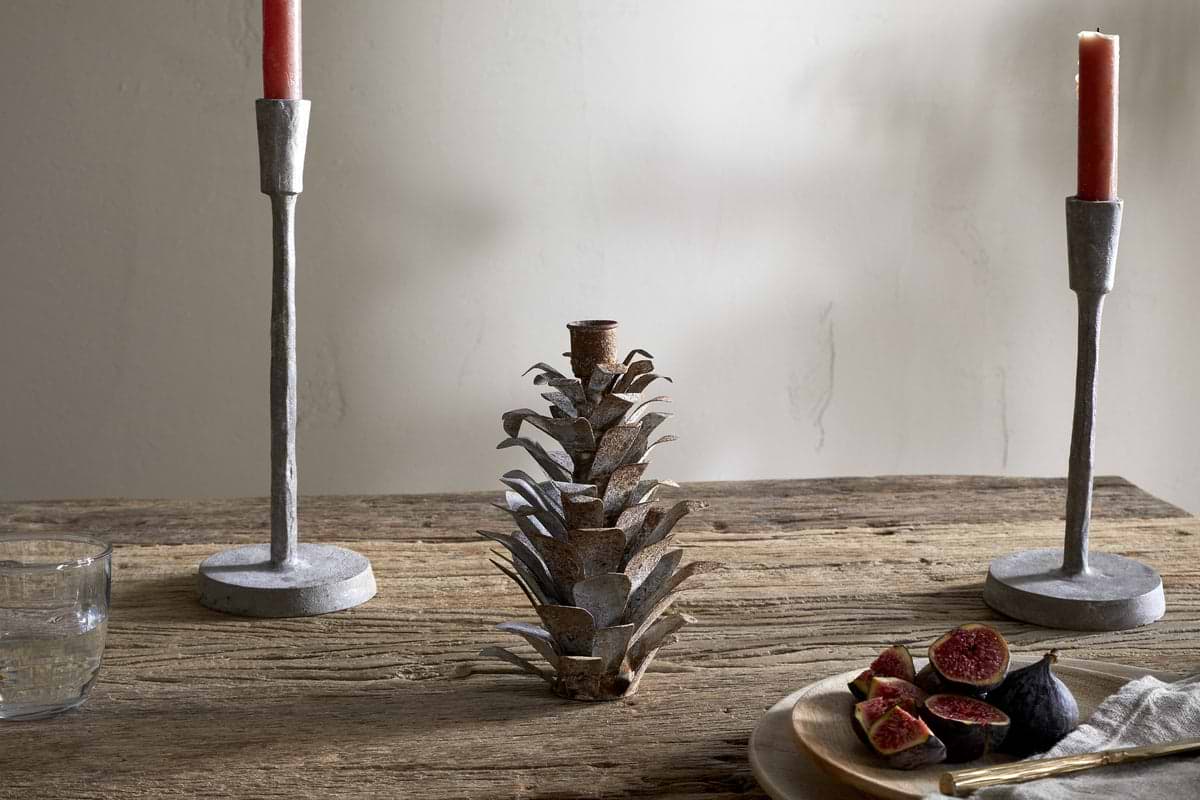 A rustic whitewashed wooden table displays two gray candlesticks with red candles, the nkuku Elagalu Pine Cone Candle Holder in zinc, a glass of water, and a wooden plate of figs against a neutral minimal background.