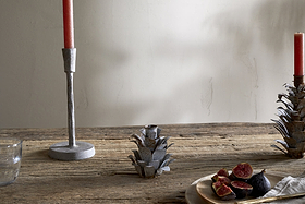 A rustic wooden table displays nkuku's Elagalu Pine Cone Candle Holders - Zinc (Set of 2), paired with a red candle, glass tumbler, metal artichoke accent, and a plate of halved figs and knife—all softly illuminated for festive charm.