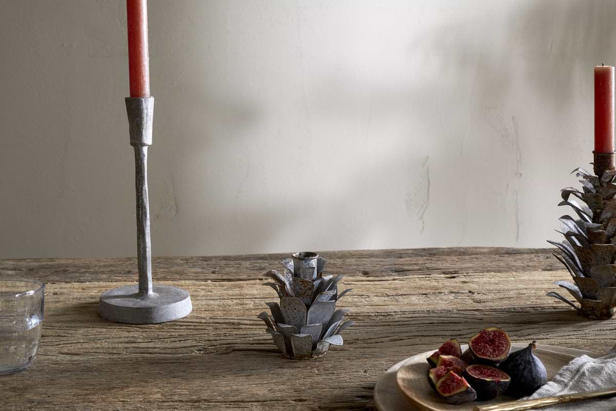A rustic wooden table displays nkuku's Elagalu Pine Cone Candle Holders - Zinc (Set of 2), paired with a red candle, glass tumbler, metal artichoke accent, and a plate of halved figs and knife—all softly illuminated for festive charm.