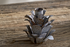 The nkuku Elagalu Pine Cone Candle Holders - Zinc (Set of 2) feature layered, metallic petals and a rustic look, making them ideal as festive tabletop decor or unique Christmas candleholders on a weathered wood surface.