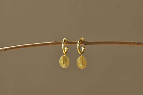 The Esta Hoop Earrings - Gold by nkuku feature oval, hammered pendants hanging from small hoops. Shown on a thin brown branch against a beige background, their minimal design adds an elegant touch. One Size.