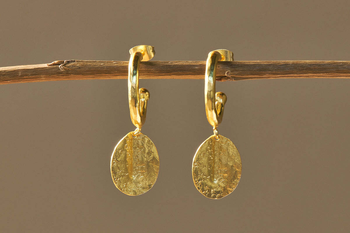 The Esta Hoop Earrings - Gold by nkuku feature oval, hammered pendants that catch the light with their polished finish. Hanging from a thin brown branch, they stand out against a simple taupe background, showcasing their handcrafted elegance.