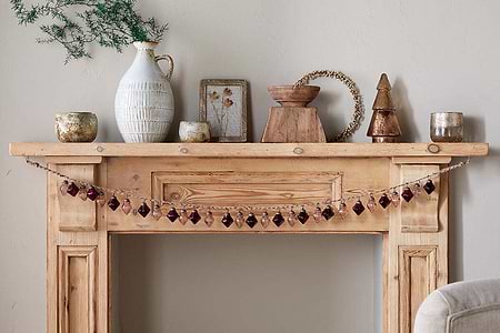 A wooden mantel displays vintage style with nkuku’s Ekiti Bauble Garland - Ruby & Copper, rustic candles, a white textured vase with greenery, framed art, a wooden bowl, beaded wreath, and metallic tree figurine against a light beige wall.